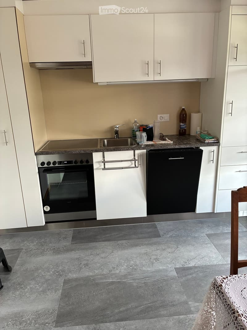 Modern kitchen with white cabinets, stainless steel oven, dishwasher, and sink. Countertop has various items including a bottle, cups, and a towel. Grey tiled floor.