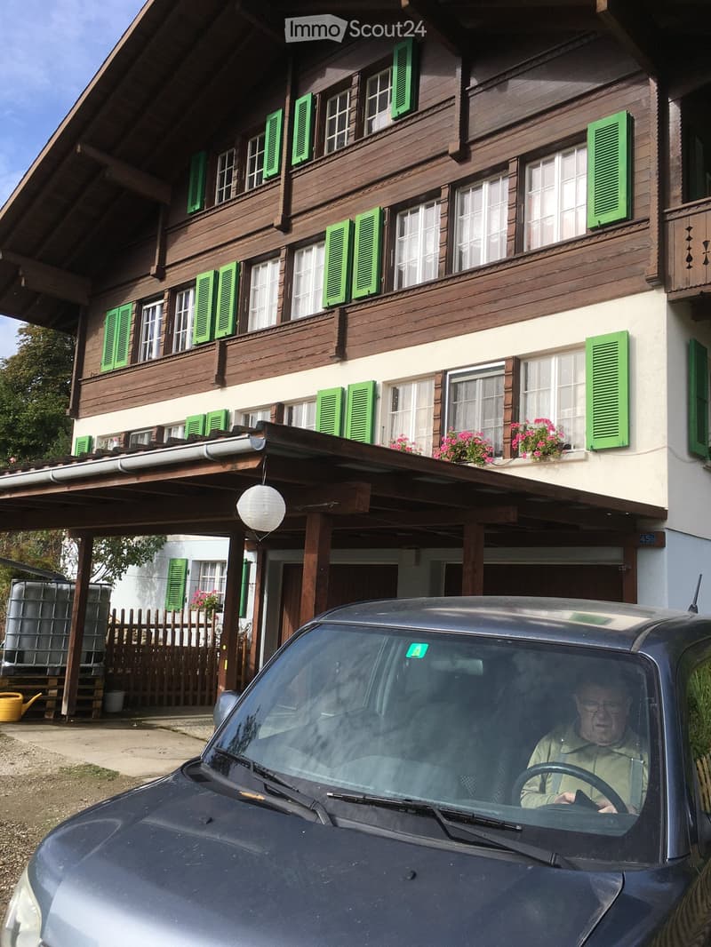 Two-story wooden house with green shutters, white-framed windows, and a covered entrance area. The house features a balcony with flower boxes and a parked car in the foreground.