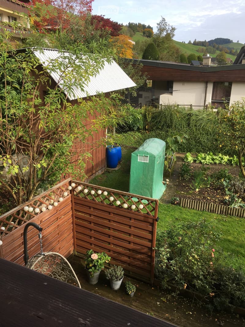 Garden area with wooden fence, potted plants, and a covered structure. The garden is surrounded by greenery and includes a small shed and a blue barrel.