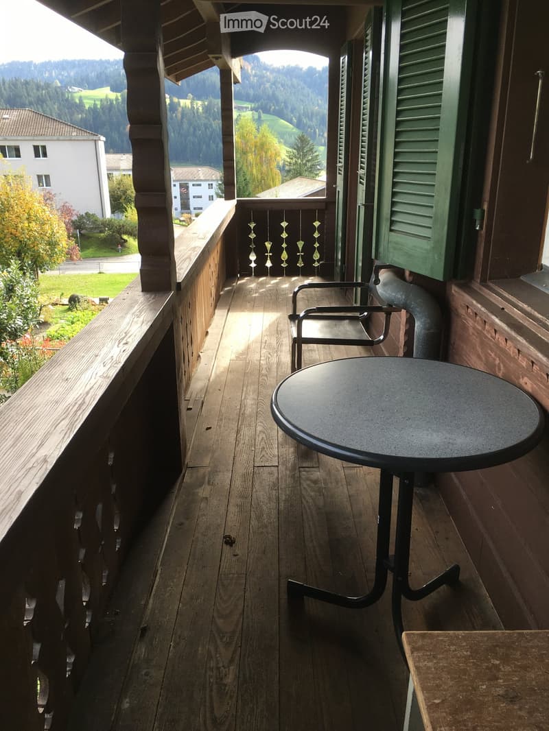 Wooden balcony with a round table and a chair, overlooking a scenic valley with trees and houses.