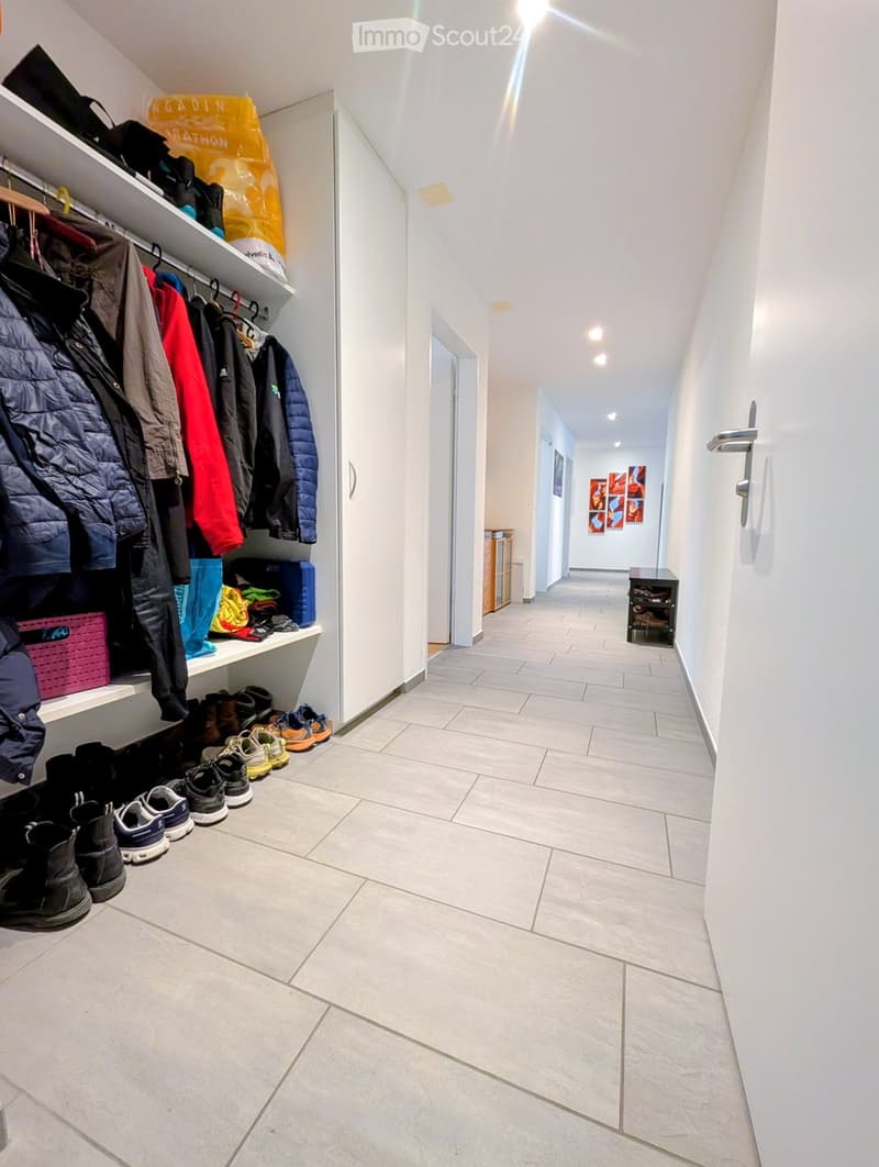 Clean, modern hallway with tiled flooring, white walls, and built-in storage unit holding coats, shoes, and bags. The hallway leads to another area with artwork on the wall.