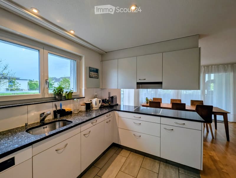 Modern kitchen with white cabinets, granite countertops, and a dining area with a table and chairs. Features include a sink, faucet, coffee maker, and small plants on the windowsill.