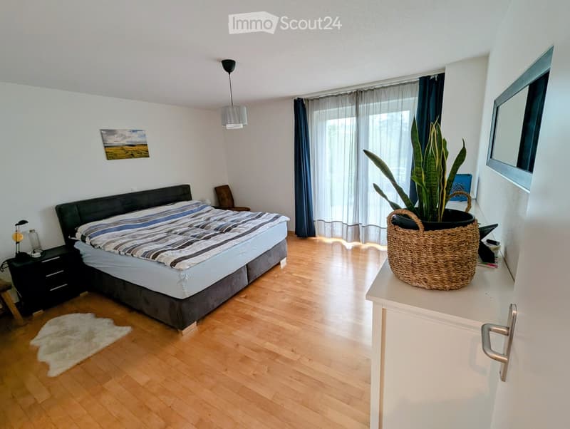 Modern bedroom with wooden flooring, a large bed with striped bedding, a nightstand with a lamp, a chair, a large potted plant on a white console table, and a mirror on the wall.