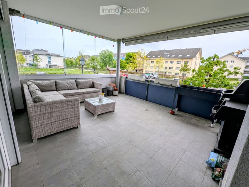 Balcony with a wicker sofa, coffee table, and grill, overlooking a residential area with trees and buildings.
