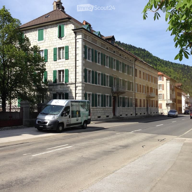 A multi-story building with green shutters and stone facade, located on a street with a parked van and other vehicles.