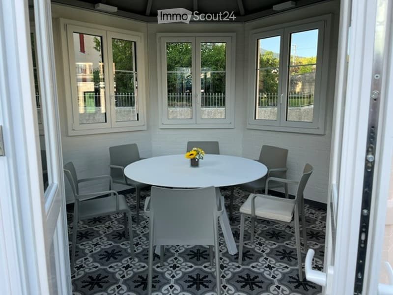 A small, enclosed balcony with white walls, multiple windows, and a round table surrounded by six chairs. The floor features a patterned tile design.