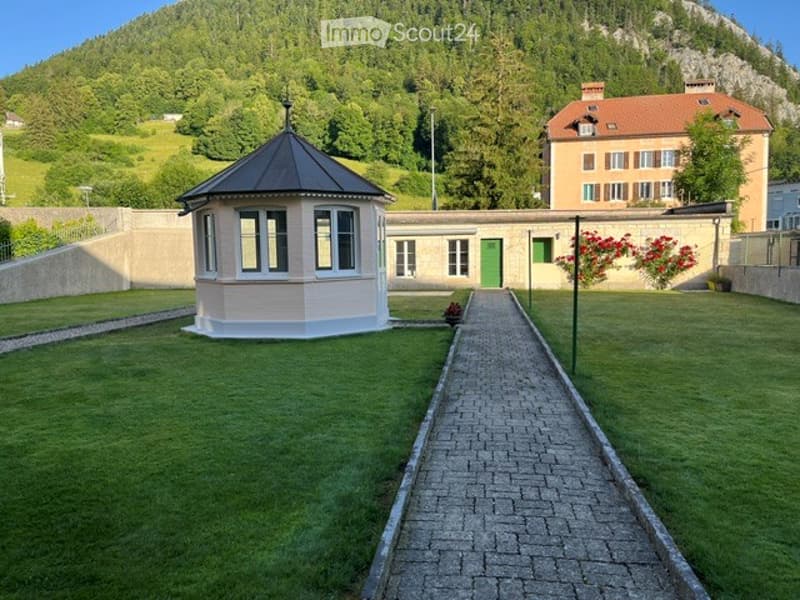 A well-maintained garden area with a small gazebo, a pathway, and a house in the background, surrounded by green hills.