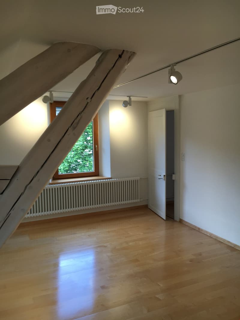 Empty attic room with wooden floors, white walls, exposed wooden beams, a window with a view of greenery, and a radiator under the window.