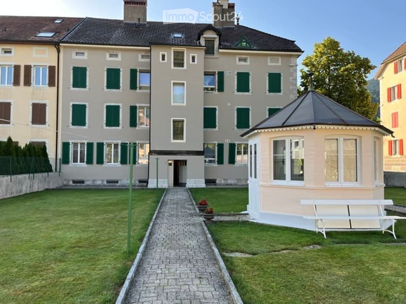 A large, multi-story building with a classic facade, green shutters, and a well-maintained lawn. A small gazebo and a white bench are visible in the foreground.