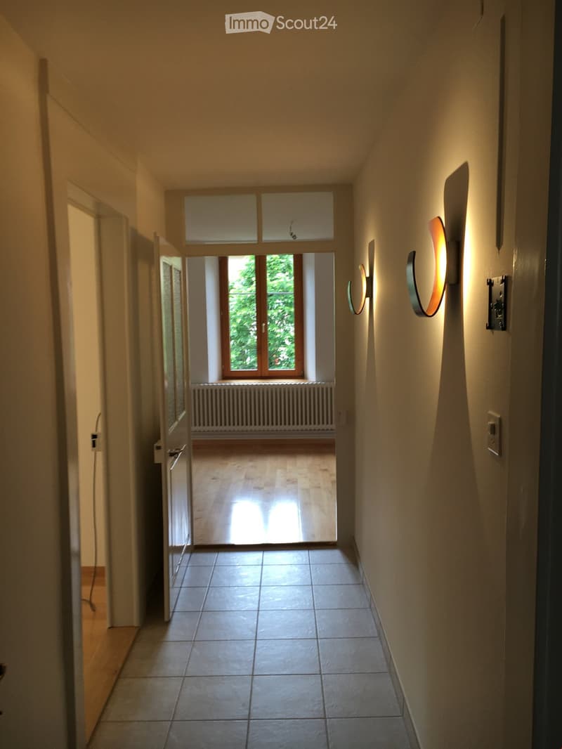 Bright hallway with tiled floor, white walls, modern wall-mounted lights, and an open doorway leading to a sunlit room with wooden flooring and a radiator.