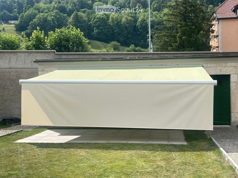 A modern outdoor patio with a large retractable awning, surrounded by a well-maintained lawn and trees, with a scenic view of a hilly landscape.