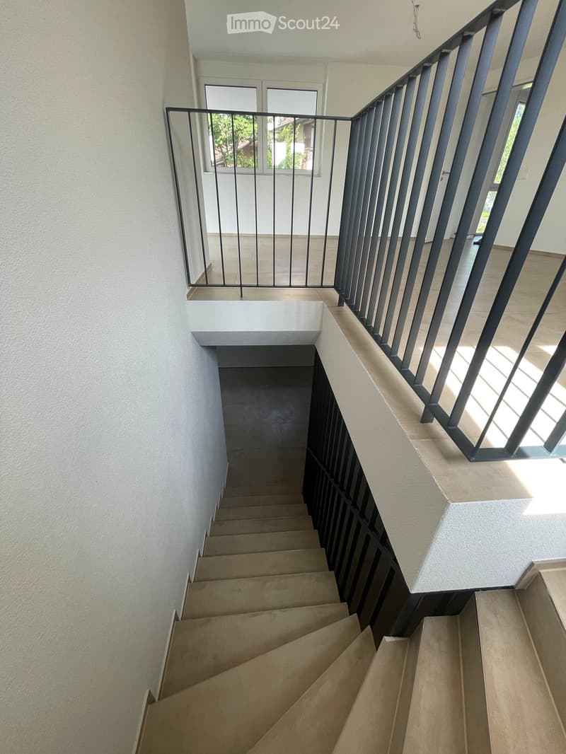 Spiral staircase with beige steps and dark metal railing, leading to an upper level with a window view of greenery.
