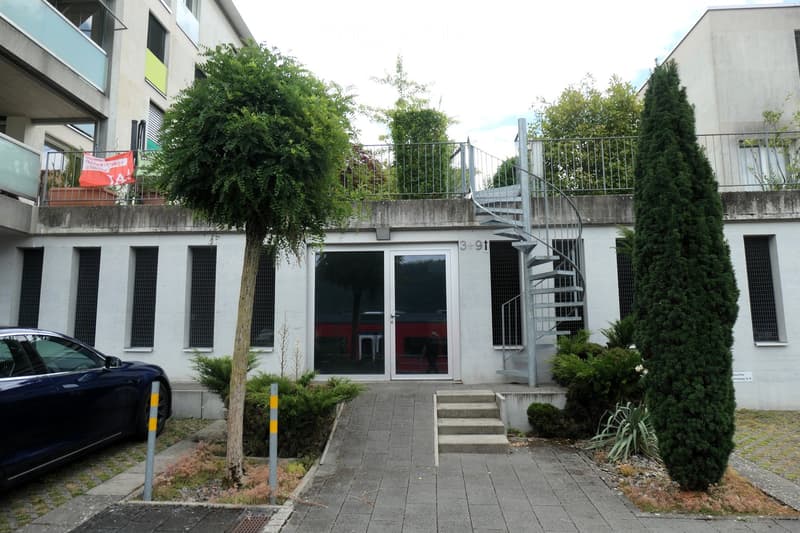 Modern building entrance with a spiral staircase leading to an upper level, surrounded by greenery and a parked car.