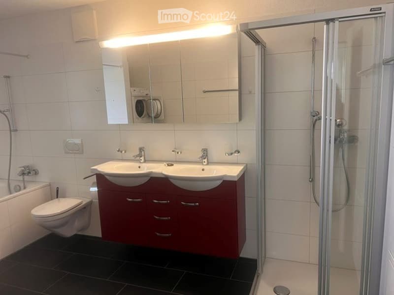Modern bathroom with double sink, large mirror, toilet, and walk-in shower. Red cabinetry and black tiled floor.
