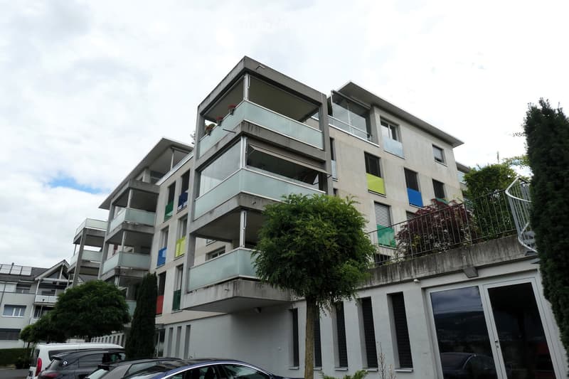 Modern multi-story apartment building with balconies, large windows, and colorful accents. Surrounded by trees and parked cars.
