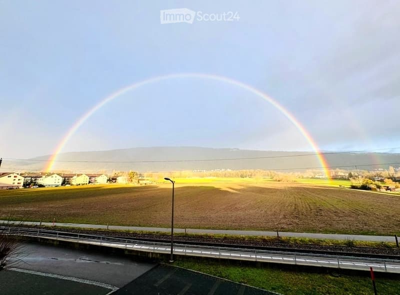 A scenic outdoor view with a double rainbow arching over a rural landscape, featuring fields, houses, and distant hills.