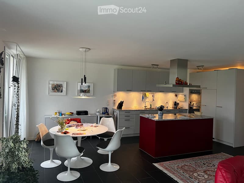 Modern kitchen with grey cabinets, a red island, a white dining table with chairs, and various appliances including an oven and a refrigerator. The space features pendant lighting and a sleek design.