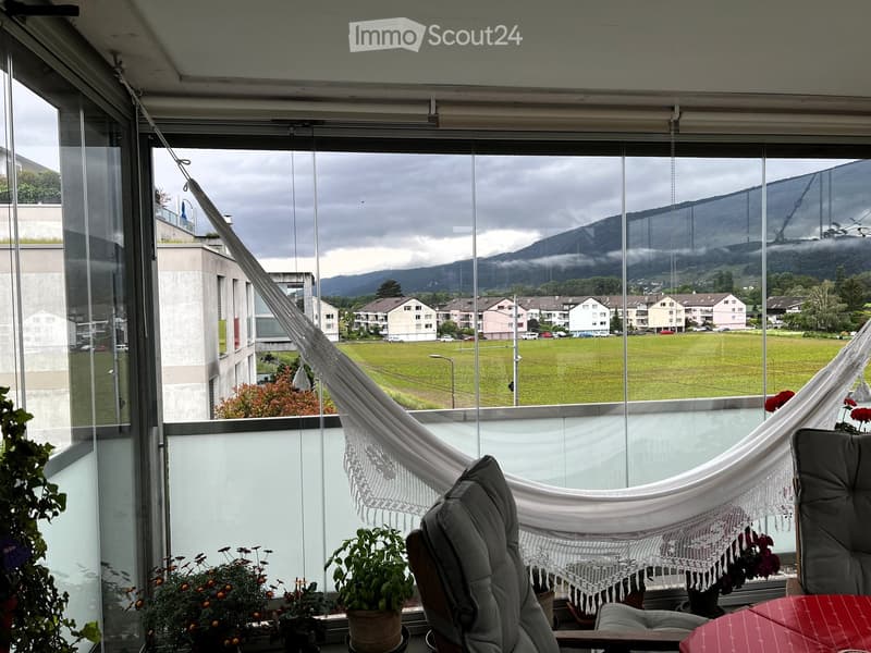 Balcony with large windows offering a scenic view of a green field, mountains, and buildings. Features a hanging chair, a small table, potted plants, and a hammock.