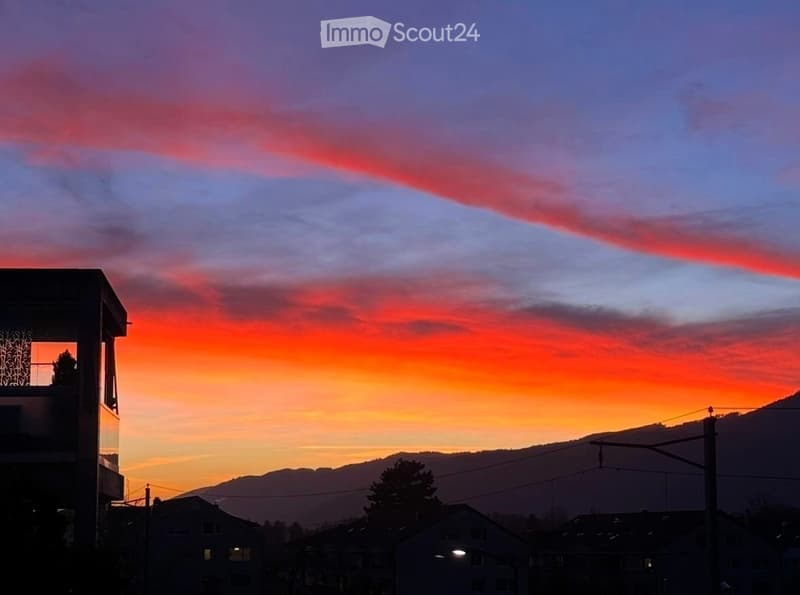 Evening view of a cityscape with a vibrant sunset sky, featuring pink and orange hues, distant mountains, and silhouettes of buildings and trees.
