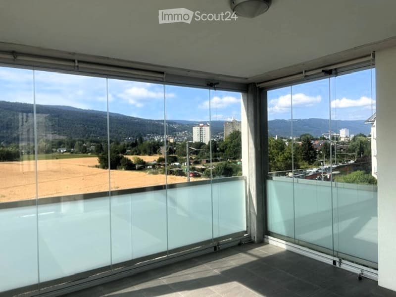 Large balcony with expansive view of a cityscape and surrounding landscape, featuring large glass sliding doors.