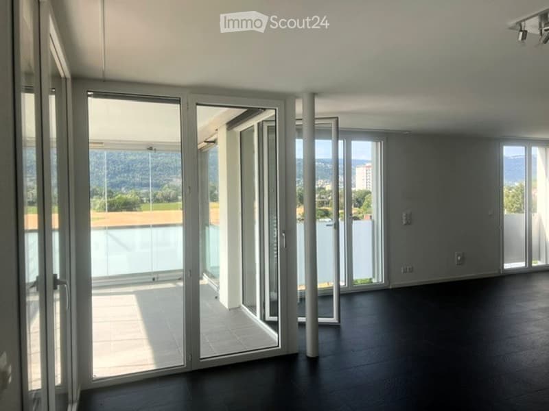 Bright, empty room with large glass doors leading to a balcony, offering a scenic view of the landscape. The room features a dark wooden floor and white walls.