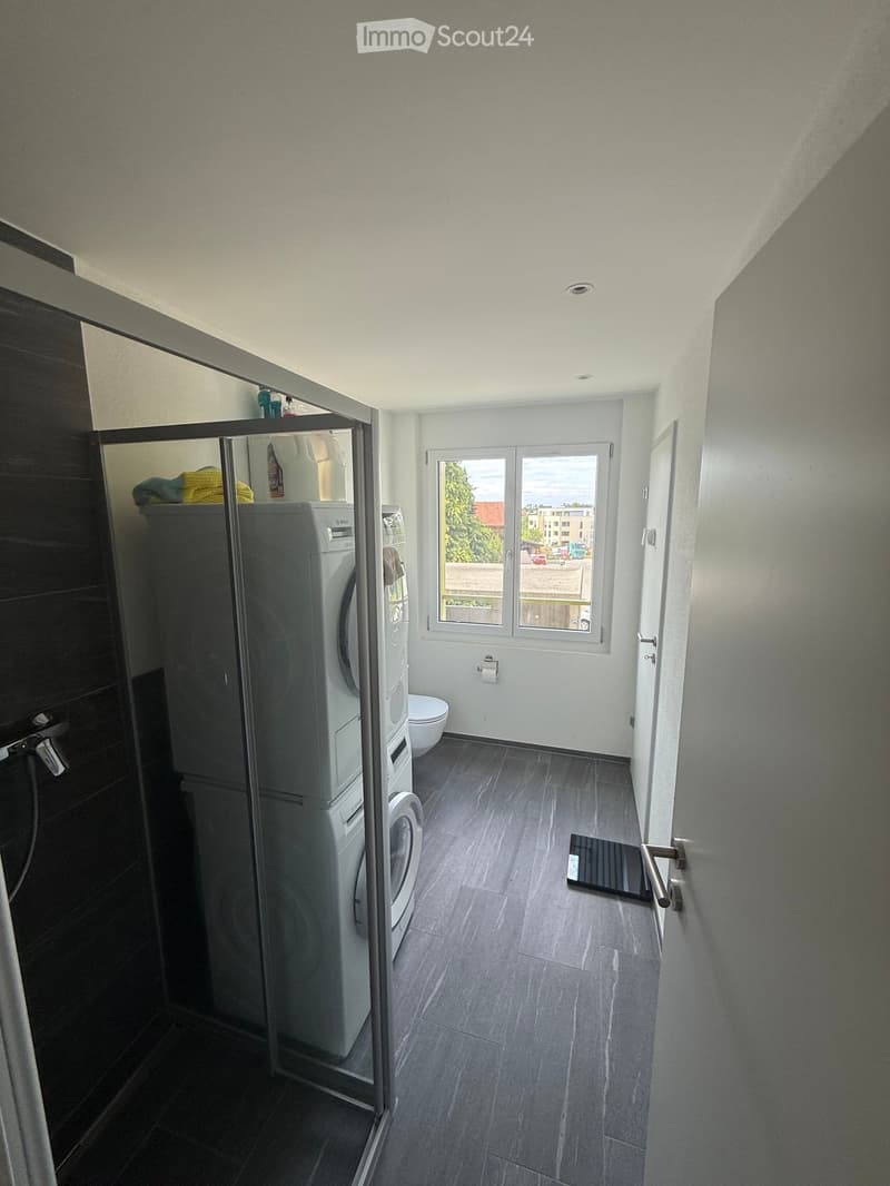 A small, modern laundry room with a washing machine, toilet, and a large window offering a view of the outside. The room features dark grey tiles on the floor and walls, with a glass shower enclosure visible on the left side.