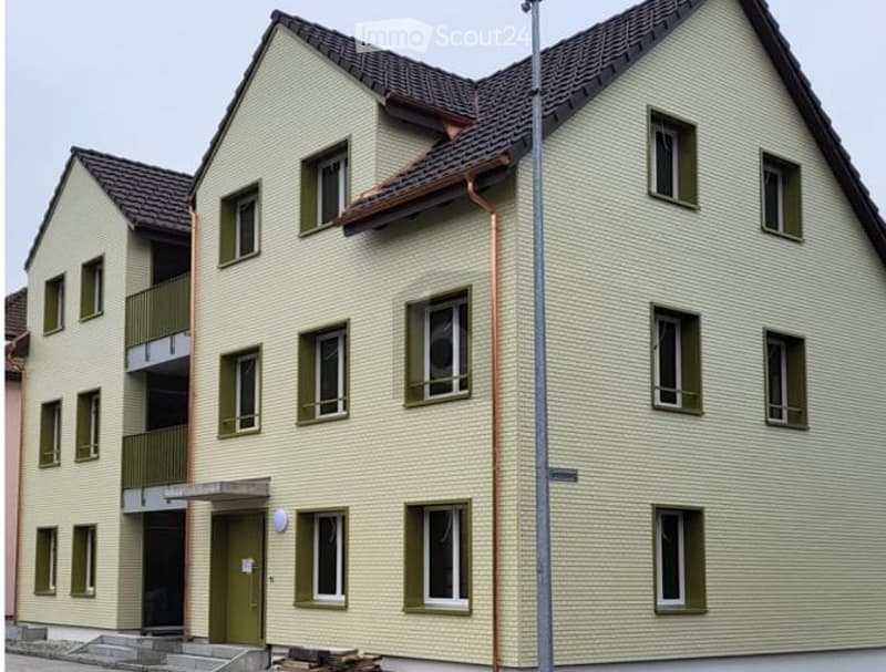 Two-story residential building with light yellow siding, green shutters, and multiple windows. Features a small balcony on the upper floor and a covered entrance area.