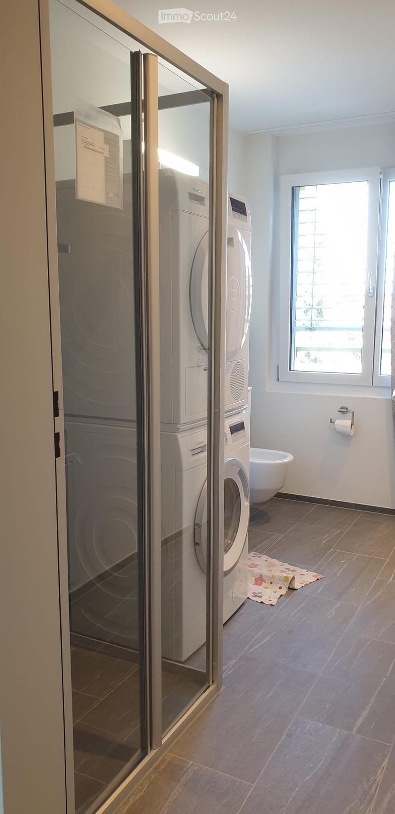 Modern bathroom with tiled floor, white walls, washing machine, dishwasher, toilet, bath, and a small rug.