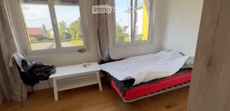 Bedroom with a single bed, a small table, and a chair near the window. The room has wooden flooring and white walls.