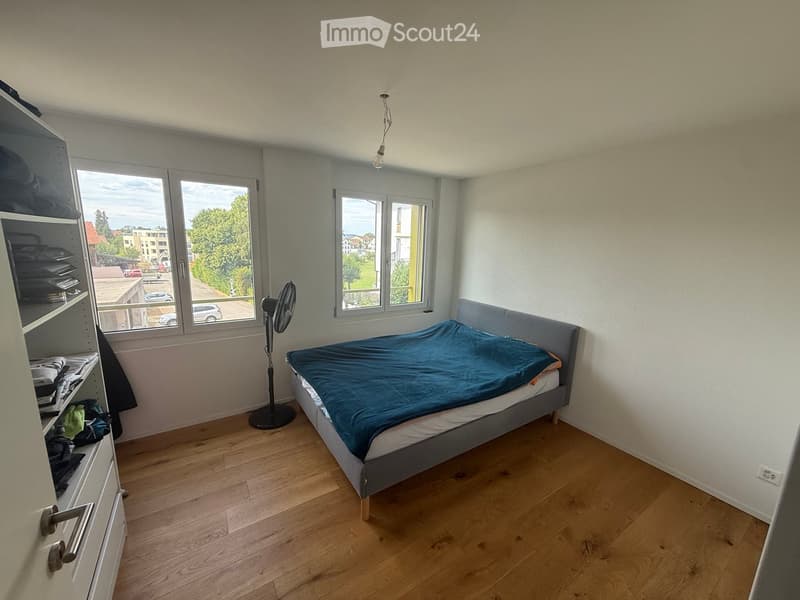 A tidy bedroom with a wooden floor, a single bed with a blue duvet, a standing fan, and a built-in wardrobe with folded clothes.