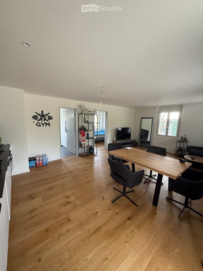 Open-plan living and dining area with wooden flooring, a large wooden dining table with chairs, a shelf with books and decor, a television, and a sofa.