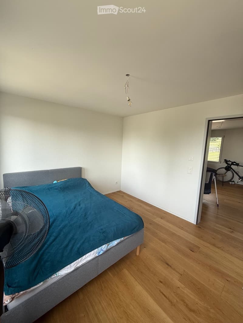 A modern bedroom with a grey headboard, blue duvet, and a standing fan on the floor. The room features light wooden flooring and white walls.
