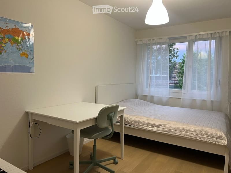 A small bedroom with a single bed, a desk, and a chair. The room features light-colored walls, wooden flooring, and a window with sheer curtains. A world map is mounted on the wall.