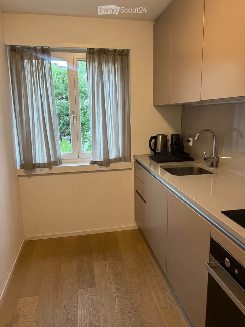 Modern kitchen with light-colored cabinets, wooden flooring, and a window with curtains. Includes a sink, faucet, oven, coffee maker, and electric kettle.