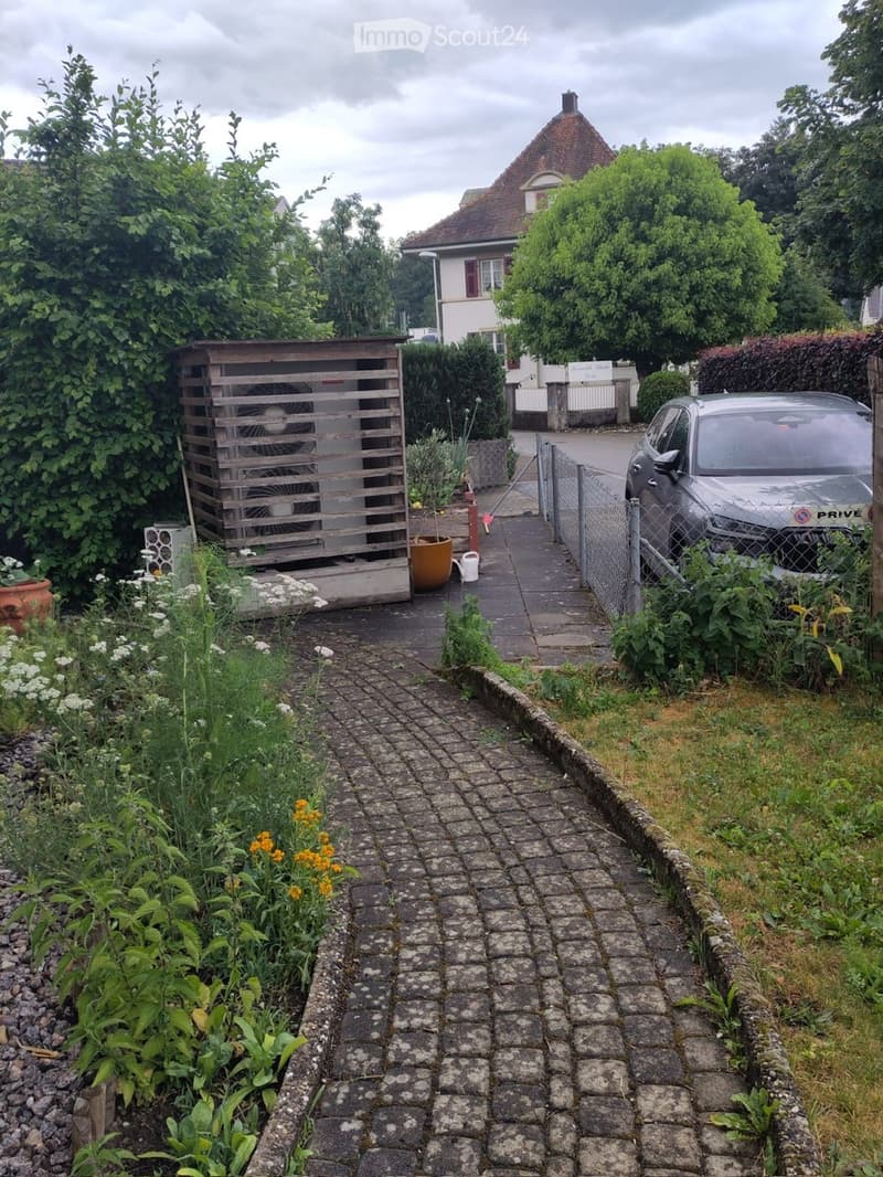 Garden pathway leading to a house, with a parked car, air conditioning unit, and various plants and flowers.