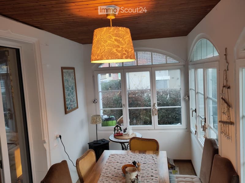 Bright living room with large windows, wooden ceiling, and a mix of seating and dining areas. Includes a sofa, dining table with chairs, a small round table, and decorative items.