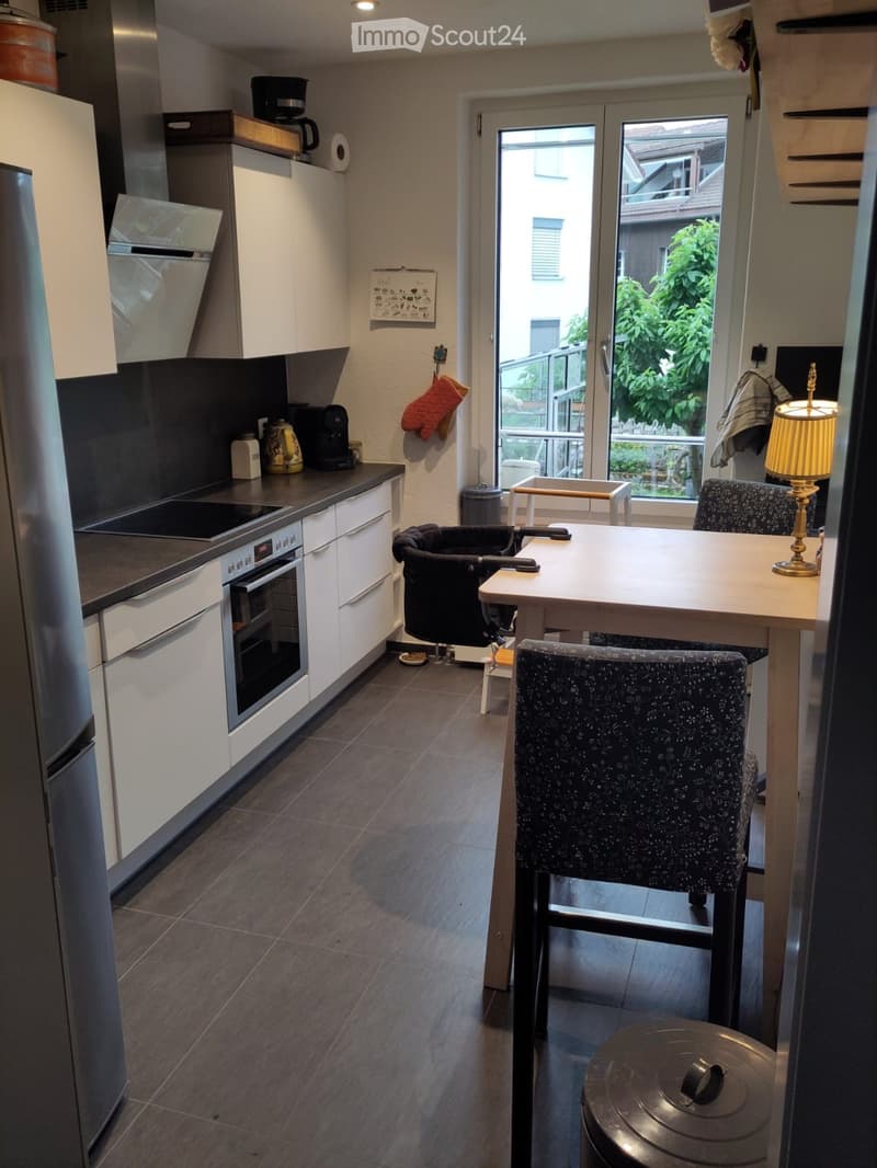 Modern kitchen with white cabinets, stainless steel appliances, and a dining area with a table and chairs. Features a large window with a view of greenery outside.