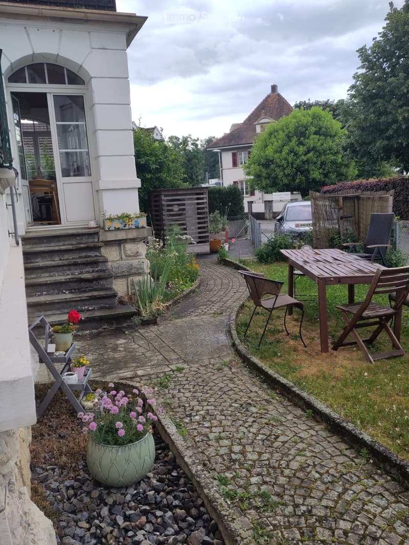 Garden area with a stone pathway, wooden table and chairs, flower pots, and a raised garden bed.