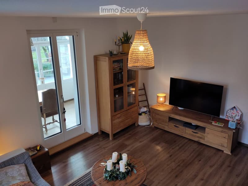 Modern living room with wooden flooring, large glass doors leading to a balcony, a sofa, coffee table with candles, TV on a wooden stand, and a wooden cabinet with glass doors.