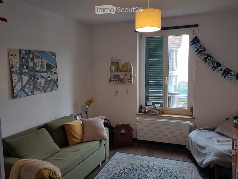 A cozy living space with a green sofa, a small bed, a shelf with books and toys, and a large window letting in natural light.