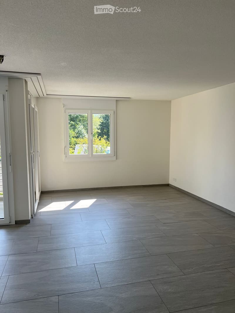 Empty room with light grey tiled floor, white walls, and a window with white frame. Sunlight streams in from the window, casting shadows on the floor.