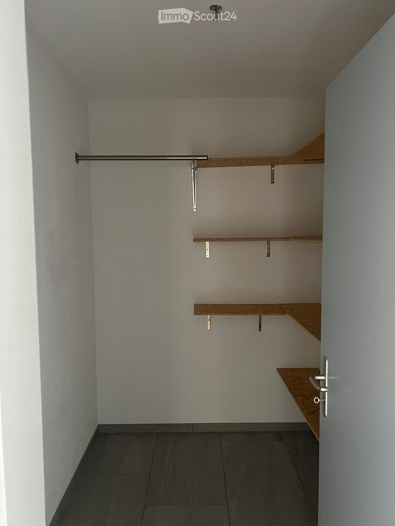 Empty closet with wooden shelves and a metal rod for hanging clothes. The room has a tiled floor and white walls.