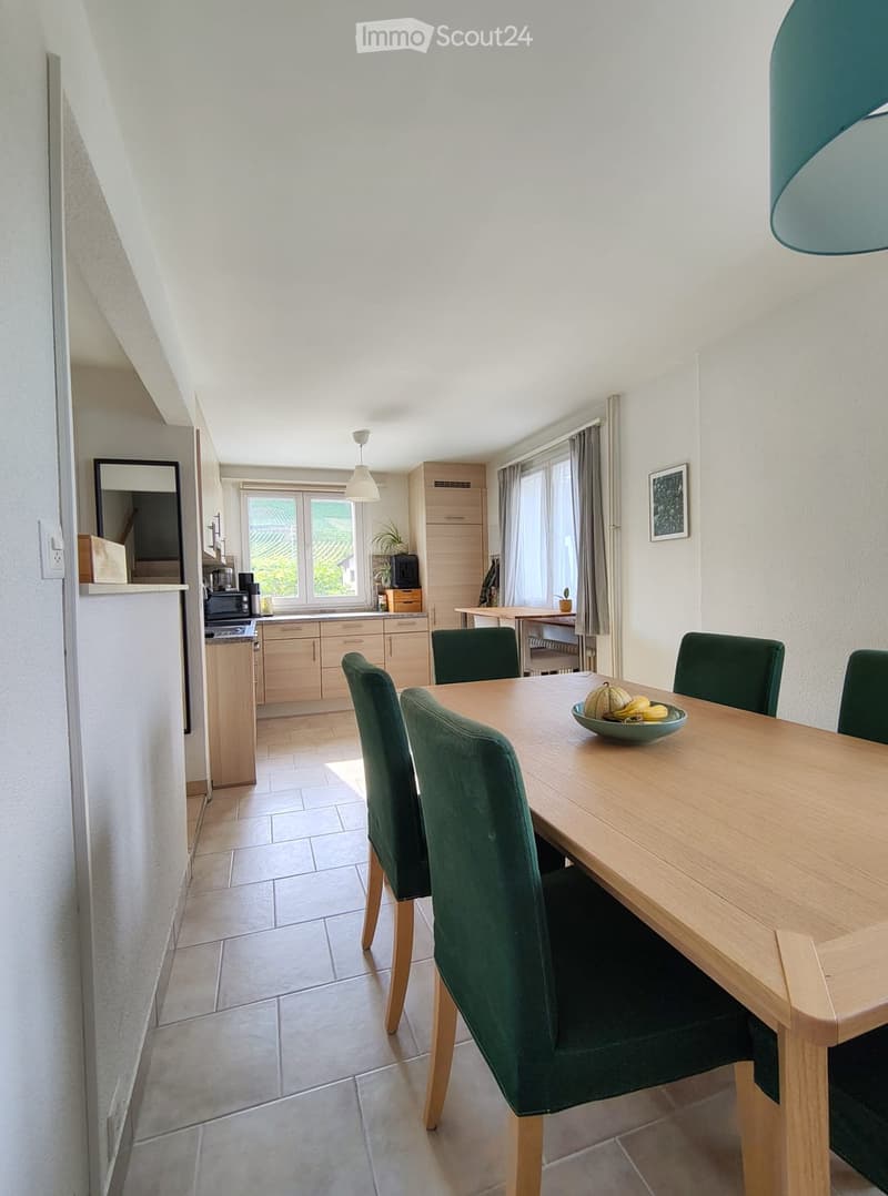 Bright kitchen and dining area with a wooden dining table, green chairs, and a bowl of fruit on the table. The kitchen features light wooden cabinets, a microwave, and a refrigerator.
