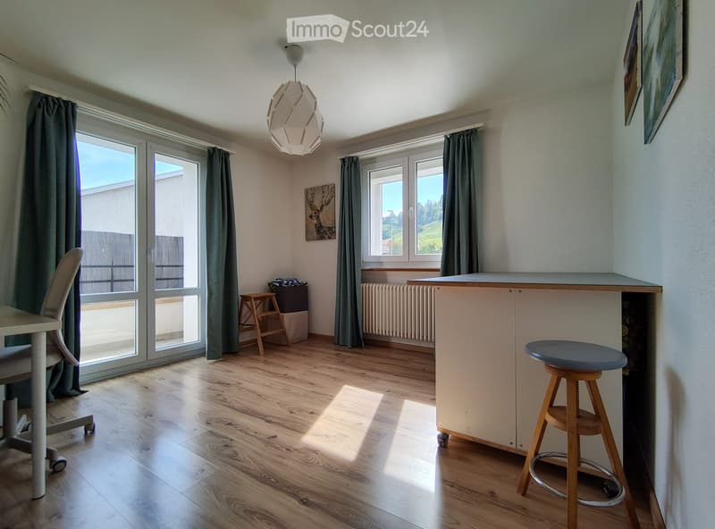 Bright room with wooden flooring, large windows, and a sliding glass door leading to a balcony. Features a white table, a chair, a small wooden stool, a bar counter with a blue stool, and a modern pendant light.