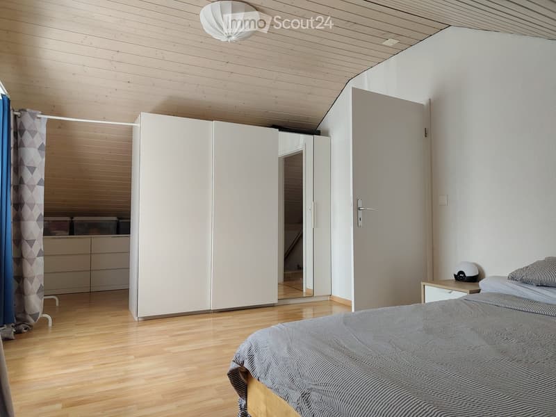 Modern bedroom with light wooden flooring, white walls, and a large sliding wardrobe with mirrored doors. Features a neatly made bed with striped bedding, a small nightstand with a lamp, and a drawer unit visible through the wardrobe doors.