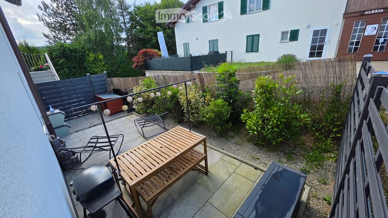 Private outdoor patio with wooden furniture, including a table and chairs, surrounded by greenery and a wooden fence. The area is enclosed and features a barbecue grill and additional seating.
