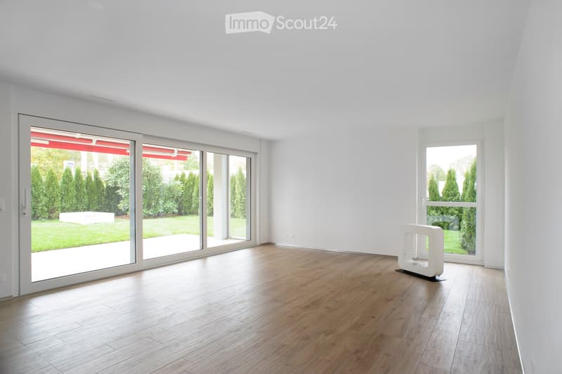 Empty room with wooden flooring, white walls, large sliding glass doors leading to a garden, and a small white decorative stand in the corner.