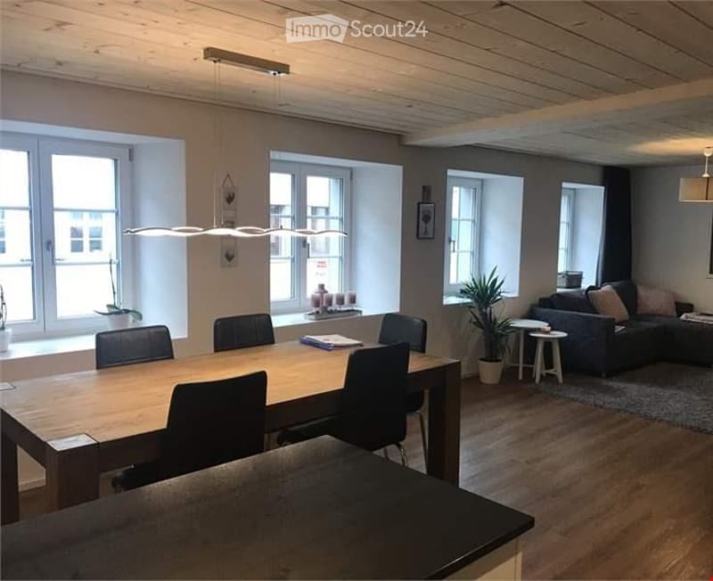 Open-plan living and dining area with wooden flooring, large windows, a dining table with chairs, a sofa, a small stool, and potted plants.