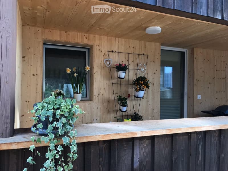 Wooden balcony with a railing, potted plants, a hanging flower rack, and a window view.