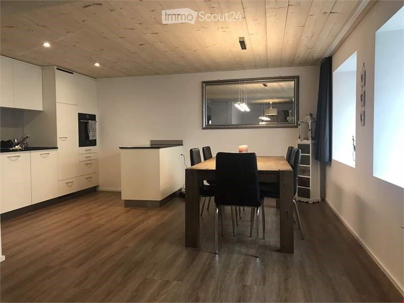 Open-plan kitchen and dining area with modern white cabinets, a wooden dining table with six chairs, a sink, oven, and a large mirror on the wall.